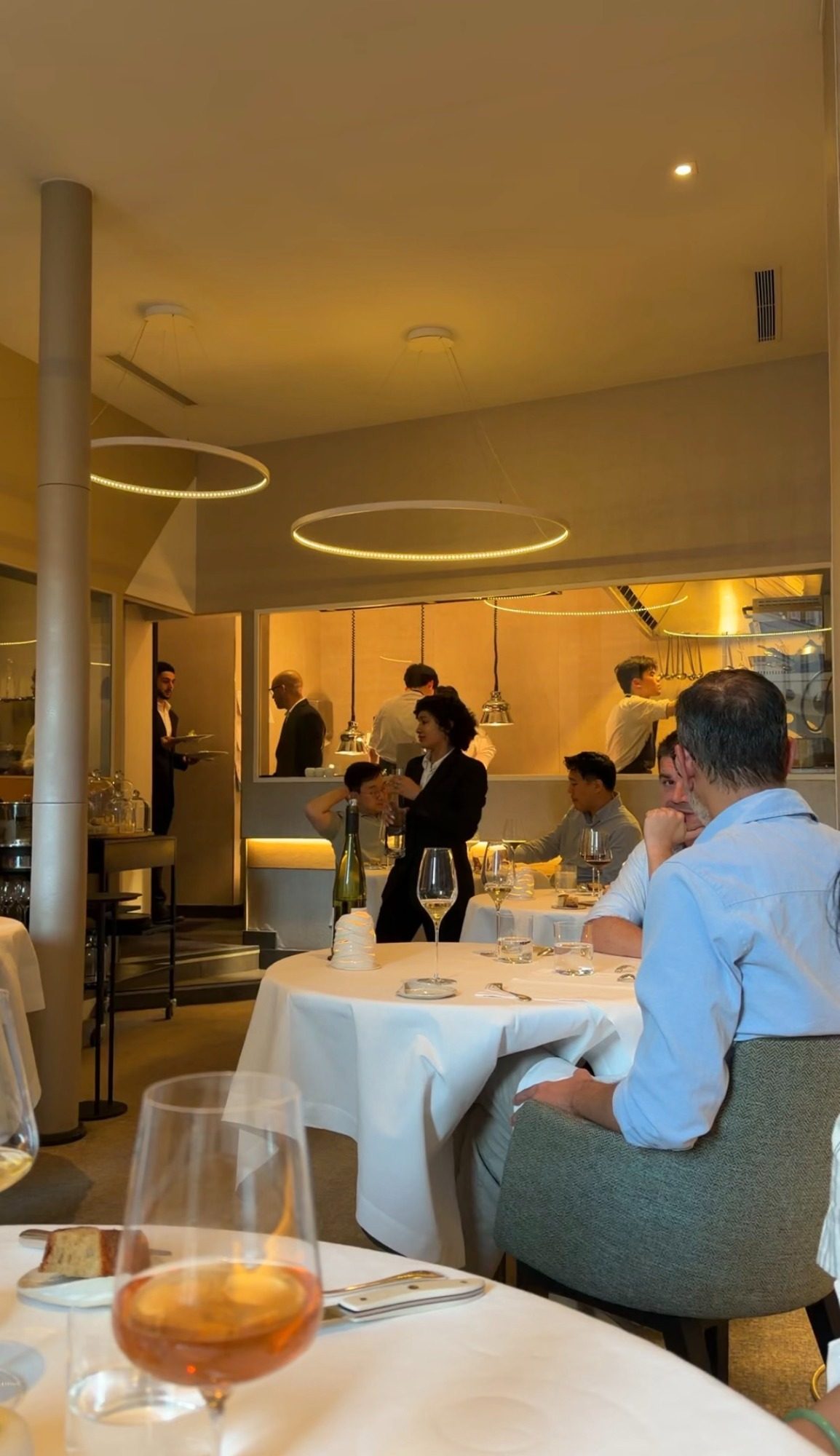 The dining room at Restaurant Alliance in Paris, with an open kitchen visible through the glass and round lights over the tables