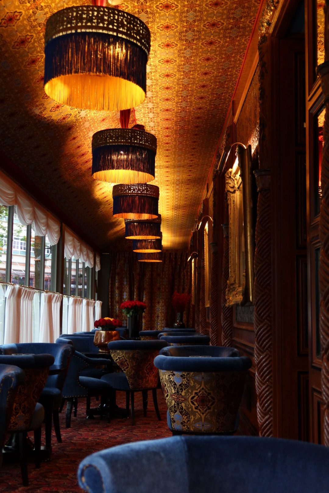 Ornate Paris cafe with blue velvet chairs and gold fringe lamps along a patterned ceiling