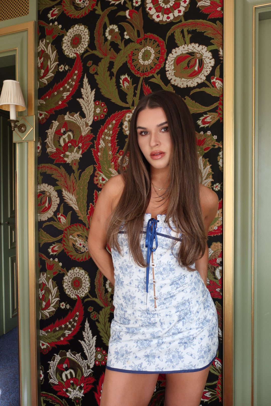 Jess in a blue and white mini dress against ornate floral wallpaper in Paris