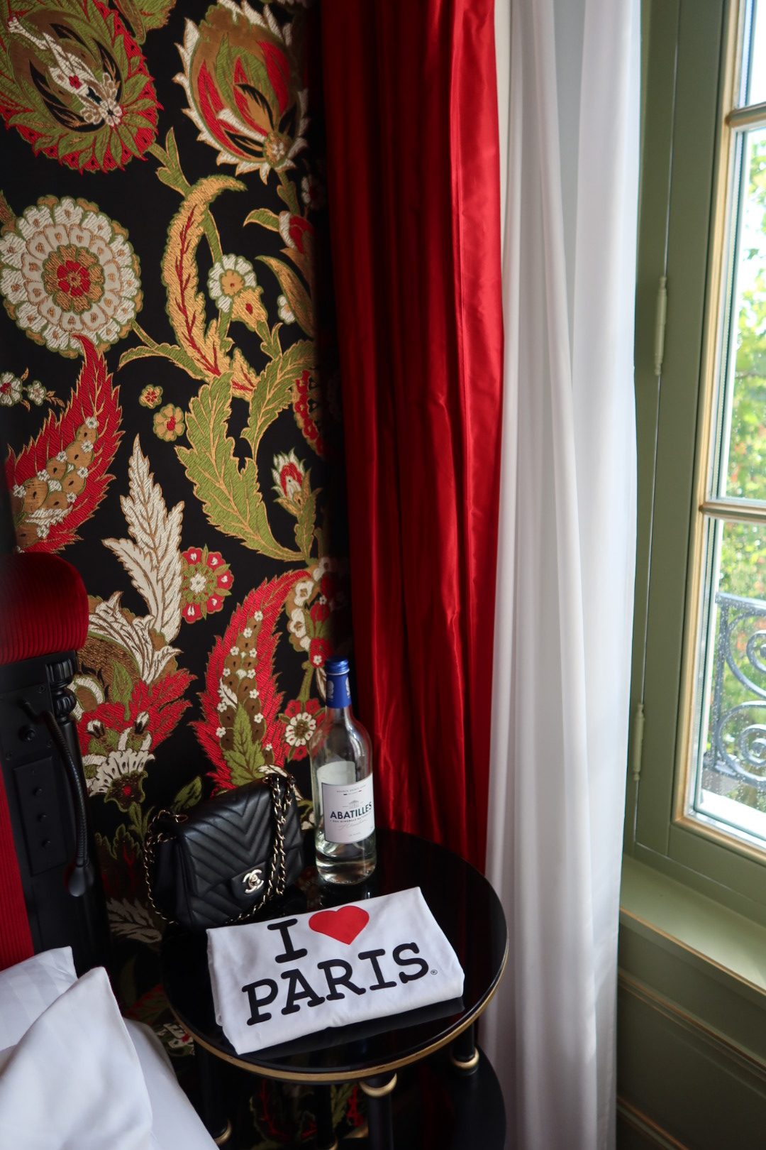 I love Paris T-shirt on a hotel table in Paris with Abatilles water, red velvet curtains, and floral wallpaper